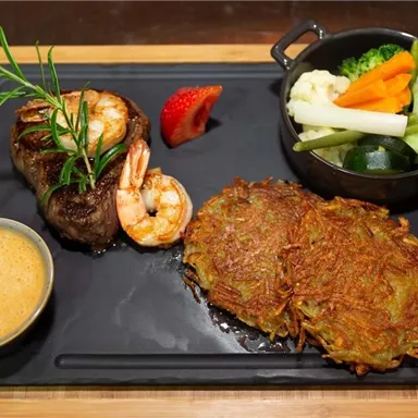 A delicious dish with beef steak, shrimp, and croquettes on a black plate. Accompanied by a portion of vegetables and a spicy sauce.