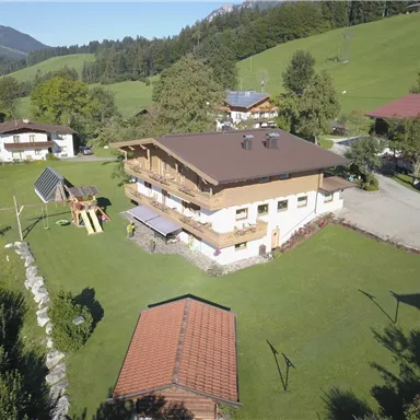 A modern, two-story house surrounded by green grass and trees. In the foreground, there is a playground, and in the background, hills and mountains.