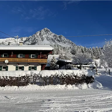 A picturesque wooden house in the snow, surrounded by snow-covered trees and mountains. The clear blue sky complements the winter landscape.