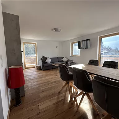 Ein modernes Wohnzimmer mit einem großen Esstisch und bequemen Stühlen. Große Fenster lassen viel Licht herein und bieten einen schönen Ausblick.