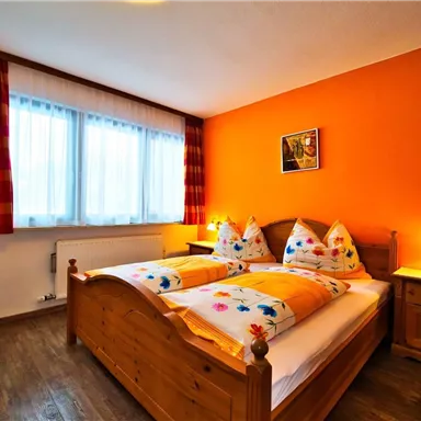 Ein gemütliches Schlafzimmer mit einem Doppelbett und bunten Kissen. Die Wände sind orange gestrichen und das Zimmer hat große Fenster mit Vorhängen.
