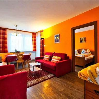 Ein gemütliches Wohnzimmer mit roten Sofas und einem Esstisch. Der Raum ist hell und freundlich, mit orangefarbenen Wänden und einem großen Fenster.