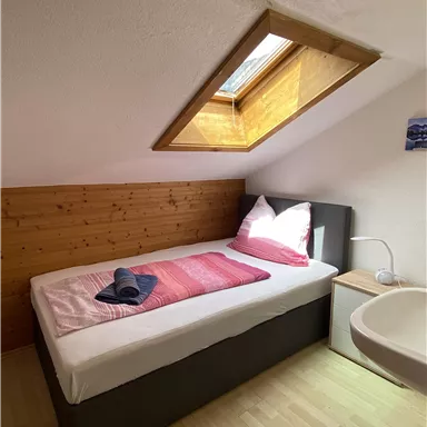 A cozy room with a single bed and a skylight. The walls are partially made of wood, and there are decorative pillows and a bedside table.