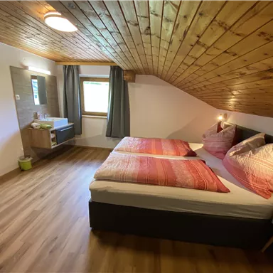 A cozy room with two single beds and wooden ceilings. There is a small desk and a window view to the outside.