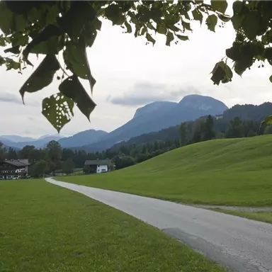 Eine ruhige Landschaft mit einer grünen Wiese und einem schmalen Weg. Im Hintergrund sind sanfte Berge und ein bewölkter Himmel zu sehen.