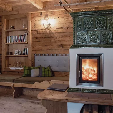 Ein gemütlicher Raum mit Holzwänden und einem klassischen Kachelofen. Eine Sitzgelegenheit mit Kissen lädt zum Entspannen ein.