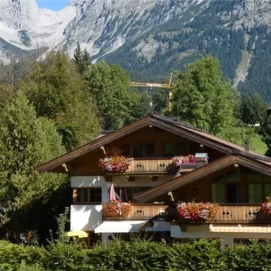 Eine malerische Berglandschaft mit saftigen Wäldern und traditionellen Chalets. Im Hintergrund sind hohe Berge und ein blauer Himmel zu sehen.