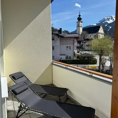 Ein Balkon mit zwei Liegen und einer schönen Aussicht auf einen kleinen Ort. Im Hintergrund sind die Berge und ein Kirchturm zu sehen.