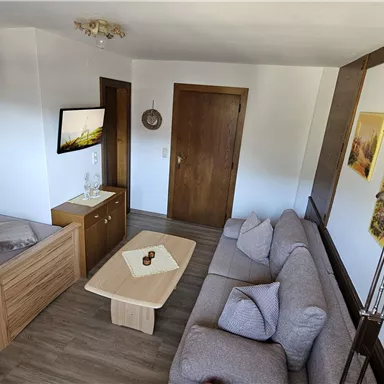 A cozy room with a single bed, a gray couch, and a wooden table. A television hangs on the wall and there are some decorative pictures.