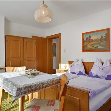 Ein gemütliches Zimmer mit zwei Betten, einem Tisch und Holz-Möbeln. Die Wände sind hell und es gibt ein Bild sowie eine Lampe auf dem Nachtisch.