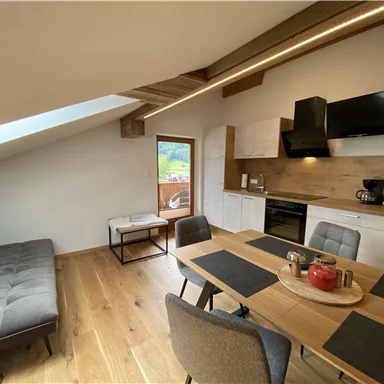 A modern, bright penthouse apartment with a cozy living area and an open kitchen. The dining table is beautifully set, and a large window provides plenty of daylight.