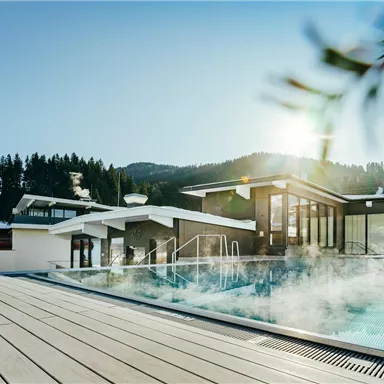 A modern pool with steam formation in a snowy environment. Trees and hills are visible in the background, while the sun shines.