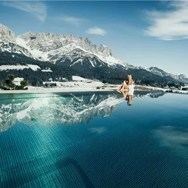 A beautiful infinity pool overlooking snow-covered mountains. In the background, a church and a clear sky can be seen.