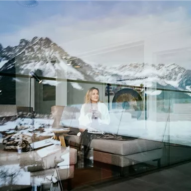 A woman sits relaxed on a sofa and looks out of a large window. In the background, snow-covered mountains and a picturesque winter landscape can be seen.