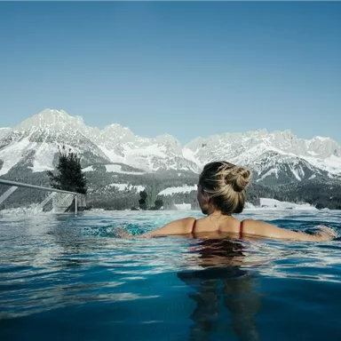 A woman relaxes in a pool with a view of snow-covered mountains. The clear sky and the mountain landscape create a tranquil atmosphere.