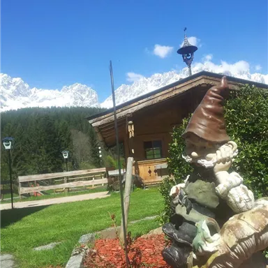 Eine kleine Hütte im Grünen mit Bergen im Hintergrund. Ein Zwerg aus Stein sitzt im Vordergrund und schaut auf die Landschaft.