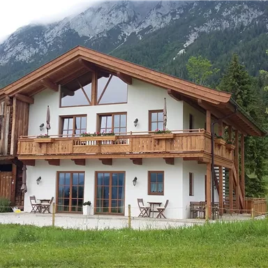 Ein modernes Landhaus mit Holzverkleidung und einem Balkon. Umgeben von einer grünen Wiese und Bergen im Hintergrund.