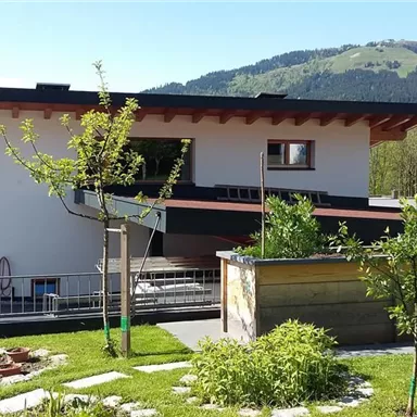 Ein modernes Haus mit einem Garten, der von kleinen Bäumen umgeben ist. Im Hintergrund sind sanfte Hügel zu sehen und der Himmel ist klar.