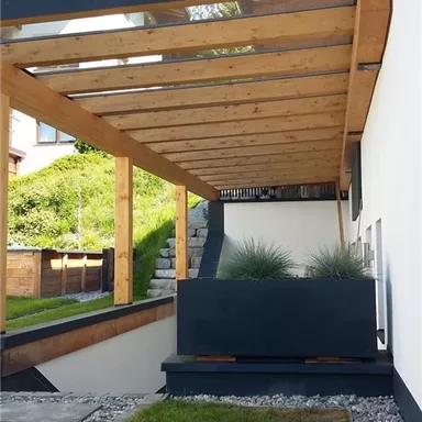Ein moderner überdachter Bereich mit Holzkonstruktion und grünem Rasen. Im Hintergrund sind einige Pflanzen und ein gepflegter Garten sichtbar.