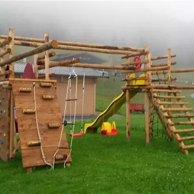 Ein Spielplatz aus Holz mit einer Rutsche, Kletterwand und Schaukeln. Die Umgebung ist grün und neblig, mit Bergen im Hintergrund.