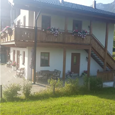 Ein charmantes Holzhaus mit zwei Etagen und einem Balkon, geschmückt mit Blumen. Der Rasen um das Haus ist grün und einladend.