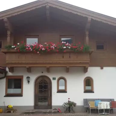 A traditional house with wooden siding and a balcony full of flowers. The entrance area is welcoming with a table and chairs in front of the front door.