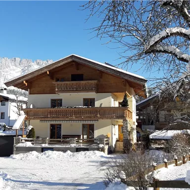 Ein gemütliches Haus im Schnee mit einem klaren blauen Himmel. Umgeben von Bergen und anderen Gebäuden, bietet es eine idyllische Winterlandschaft.
