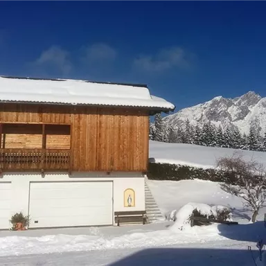 A rustic wooden house covered in snow, surrounded by picturesque mountains. The sky is clear and blue.