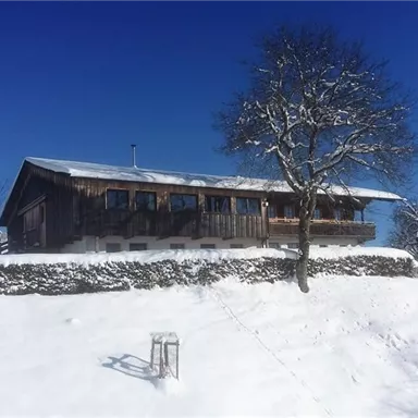 A cozy wooden house sits on a snow-covered hill. The clear blue sky and the snow-capped mountains in the background give the scene a winter tranquility.