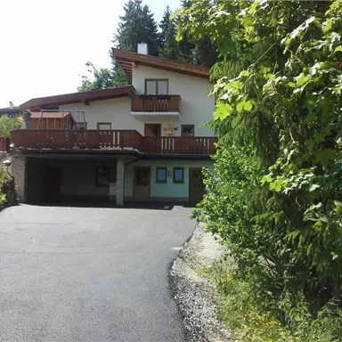 Ein modernes Haus mit einem Balkon und einer Auffahrt, umgeben von Bäumen. Die Aufnahme zeigt eine ruhige, grüne Umgebung.