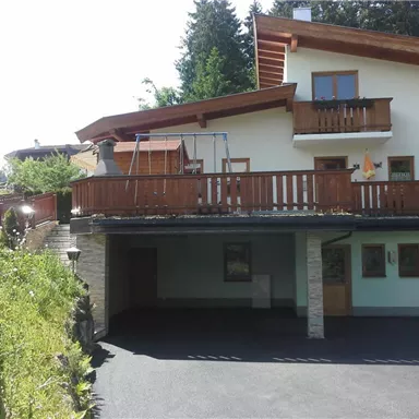 Ein modernes Haus mit einem großen Balkon und einer Terrasse. Die Umgebung ist grün und es gibt eine Auffahrt.