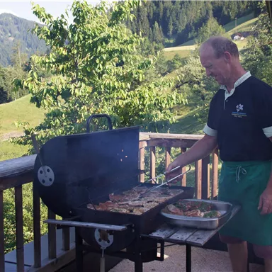 Ein Mann grillt auf einer Terrasse mit Blick auf grüne Hügel. Rauch steigt vom Grill auf und es ist ein sonniger Tag.