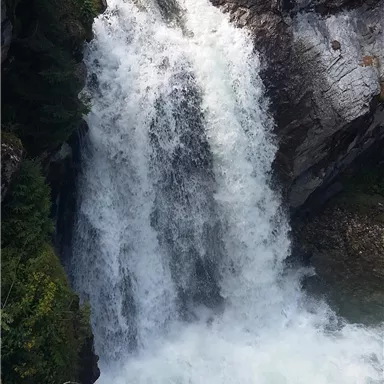 Ein rauschender Wasserfall stürzt über Felsen in ein klares Wasserbecken. Umgeben von grüner Vegetation bietet die Szene eine friedliche Naturkulisse.