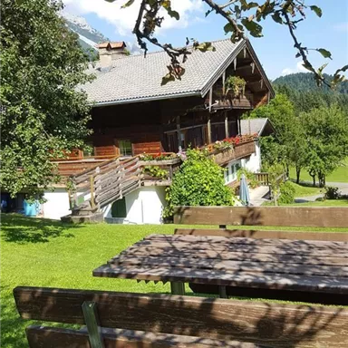 A traditional chalet surrounded by green grass and trees. In the foreground, there is a wooden bench outdoors.