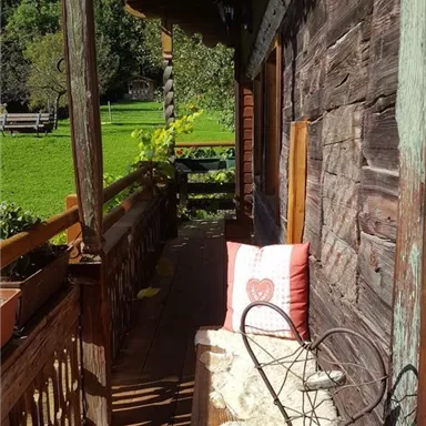A cozy balcony with wood-paneled walls and a seating area. On the balcony are cushions and a blanket, surrounded by plants.