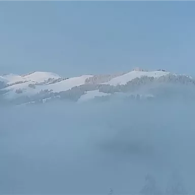 Eine verschneite Landschaft mit Hügeln, die teilweise in Nebel gehüllt sind. Der Himmel ist klar und blau.