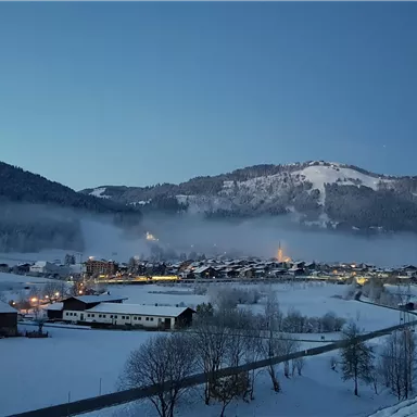 A snowy landscape at dawn with gentle hills and a small village. The lights of the houses glow softly through the fog.