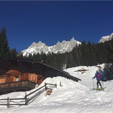 Eine idyllische Berghütte im Schnee, umgeben von hohen Tannen und majestätischen Gipfeln. Eine Person bewegt sich auf Skiern durch die verschneite Landschaft.
