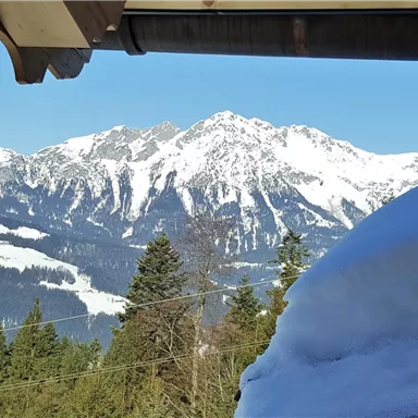 Eine herrliche Berglandschaft mit schneebedeckten Gipfeln und einem klaren blauen Himmel. Im Vordergrund sind schneebedeckte Dächer und Bäume zu sehen.