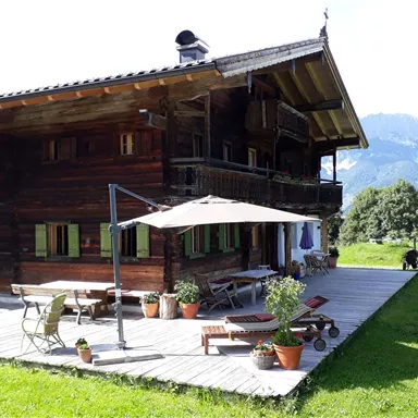 A rustic wooden house with a large deck and several pots with plants. In the background, gentle hills and mountains are visible.
