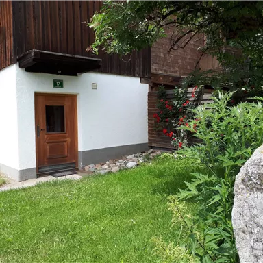 A cozy house with a white facade and a wooden porch. It is surrounded by green grass and colorful flowers.