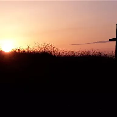 Ein Sonnenuntergang über einer Hügelkante mit hohem Gras. Im Vordergrund steht ein Kreuz silhouettiert gegen den bunten Himmel.
