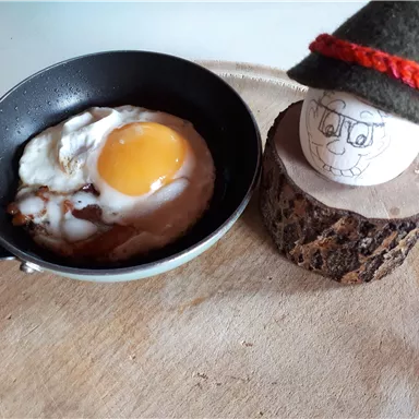 A fried egg in a small pan, served on a wooden coaster. Next to the egg is a painted eggman with a green hat.