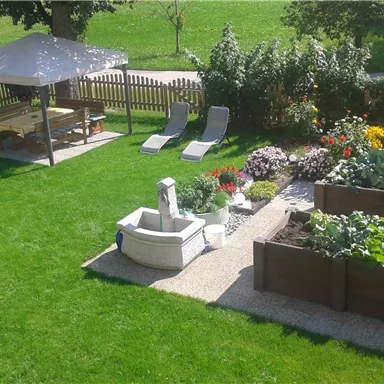 Ein gepflegter Garten mit einem Pavillon, Liegen und bunten Blumen. Hochbeete mit Gemüse und Pflanzen sind ebenfalls zu sehen.