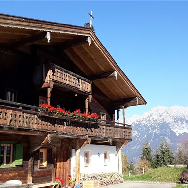 A traditional wooden house with balconies and colorful flower boxes. In the background, majestic mountains and a blue sky can be seen.