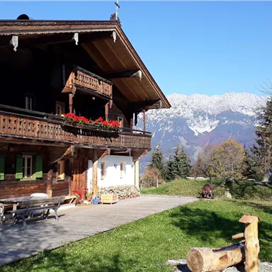 A traditional wooden house with a balcony and colorful flowers. In the background, snow-covered mountains and a clear blue sky can be seen.