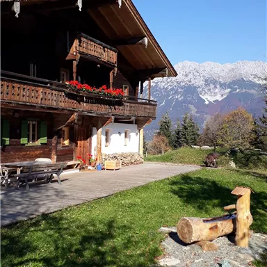A traditional chalet with blooming balcony plants and a view of the mountains. The sky is clear and blue, perfect for a relaxing day in nature.