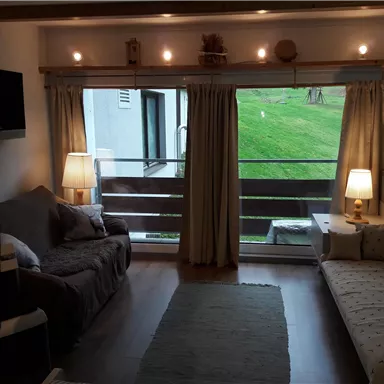 A cozy living space with two sofas, illuminated by soft lamps. Large windows provide a view of a green landscape.