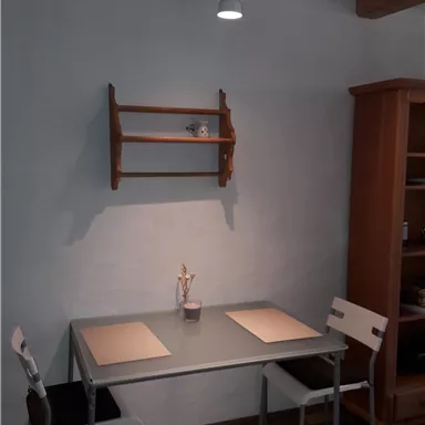 A simple dining room with a table and two chairs. There is a small wooden shelf on the wall.