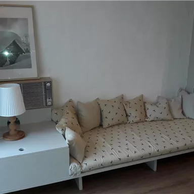 A cozy living room with a long sofa decorated with cushions. Next to the sofa is a side table with a lamp and an air conditioning unit.